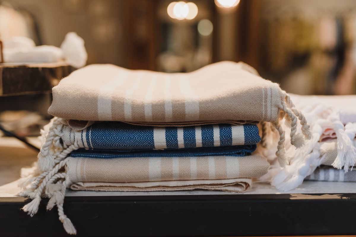 Folded bath towels in cream and warm grey stacked on a wooden bathroom shelf beside a rolled hand towel and a small green plant