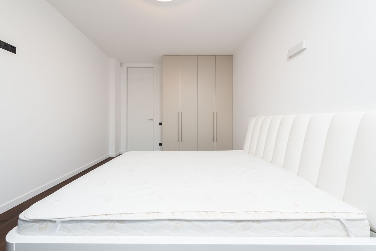 A bare white mattress on a wooden bed frame in a plain bedroom, photographed from the side to show its full length