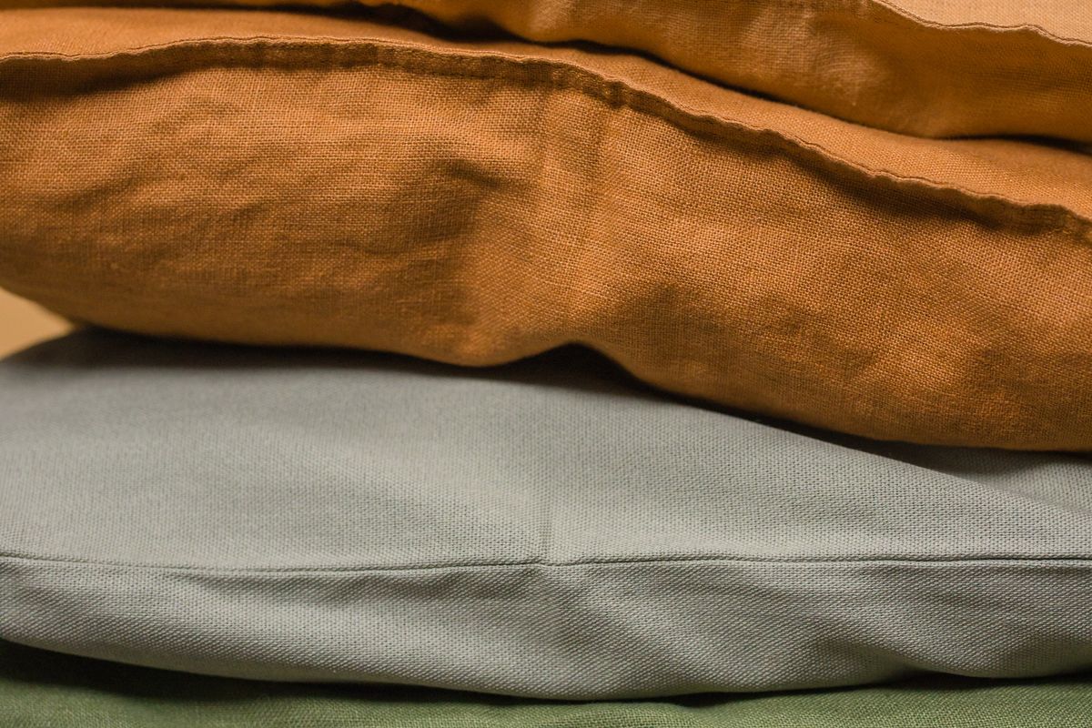 Three pillows of different thicknesses stacked on a made bed in a bright bedroom, with one older flattened pillow resting against a fresh plump one for comparison