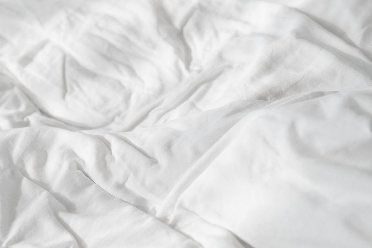 A neatly made bed with rumpled natural linen sheets in warm oat tones beside folded white cotton sheets, showing the texture difference between the two fabrics in soft daylight