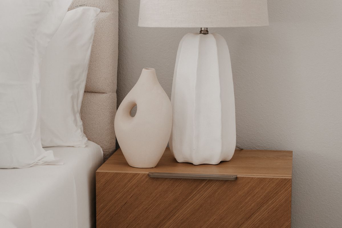 A minimalist wooden nightstand beside a bed holding a small ceramic vase and a warm table lamp in soft evening light