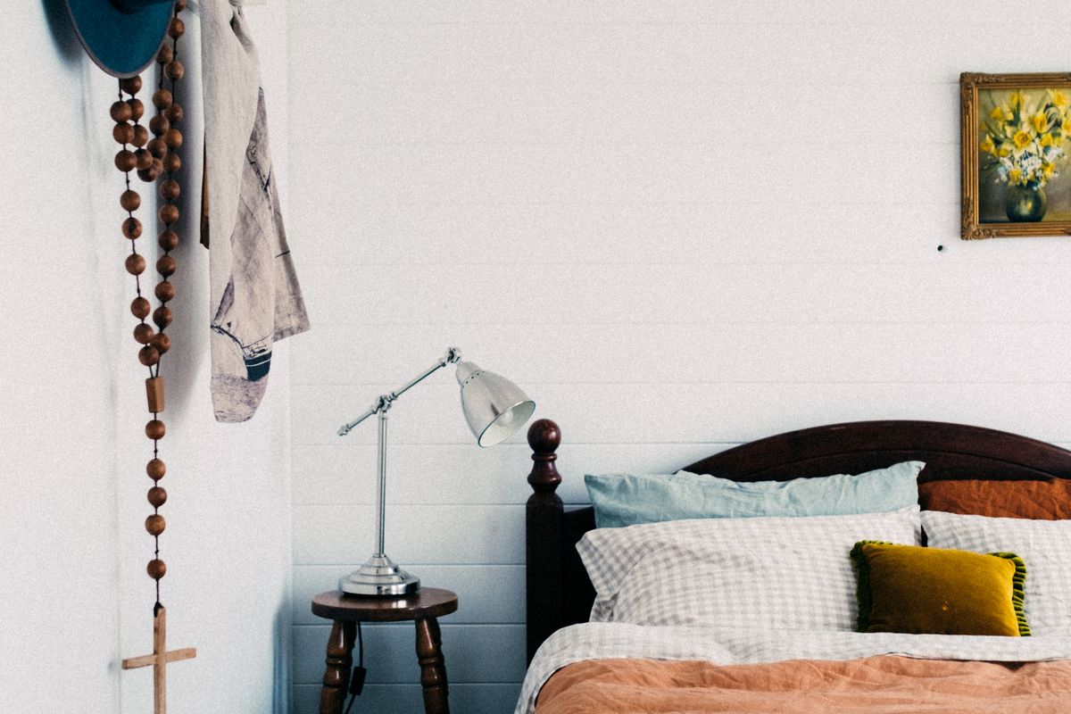 A small, neatly arranged bedroom with a bed against the wall, a simple wooden chair, and daylight from a side window